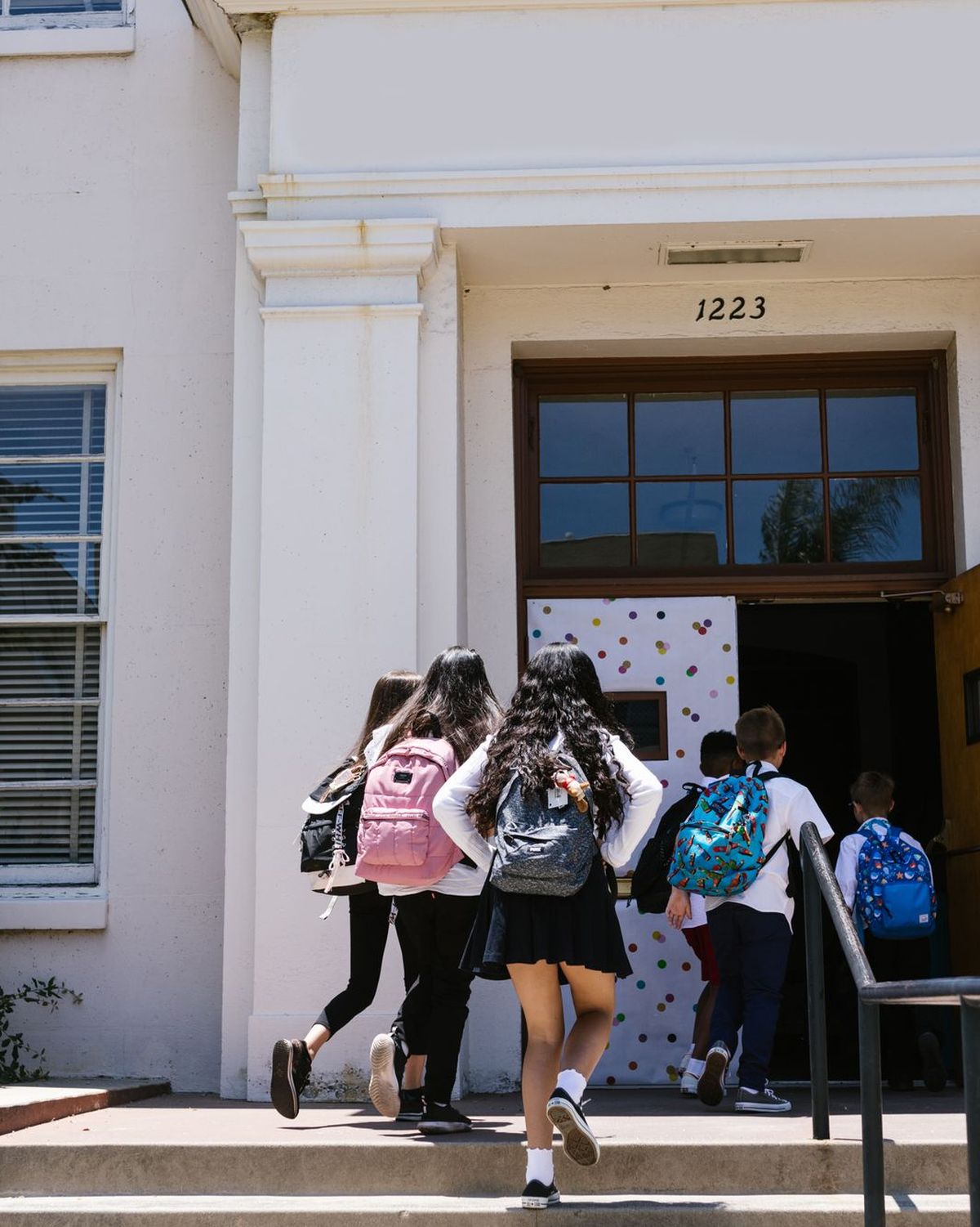 Students of different ages with backpacks walking into a school, seen from behind